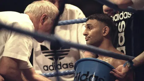 Getty Images A boxer sits in the corner of a boxing ring, being attended to by his team - one holding an Adidas-branded bucket and others dressed in Adidas gear - offering support and guidance between rounds.
