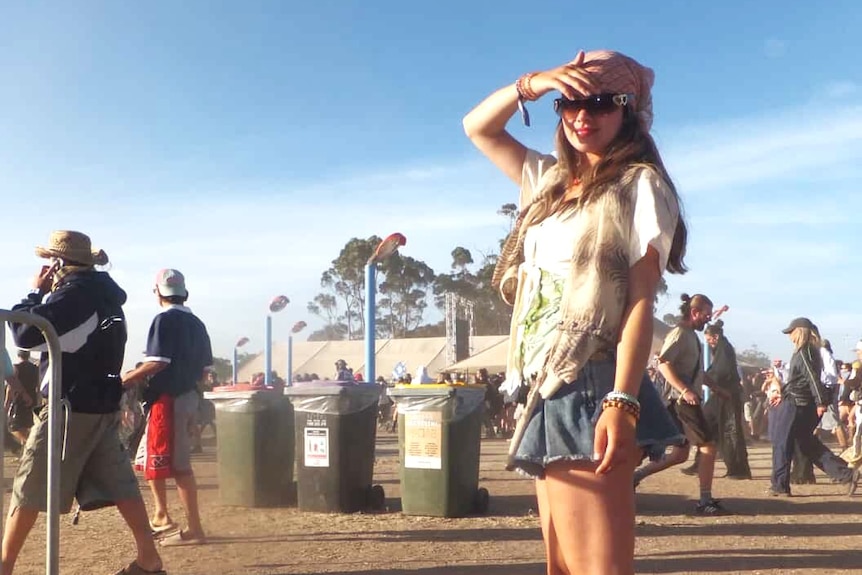 a girl standing in a dusty festival field, she is wearing sunglasses, there are large groups behind her