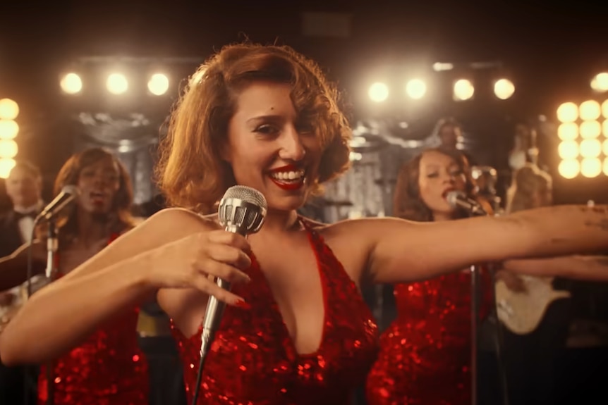 Raye stands and smiles in a red sequin dress with a vintage microphone in her music video
