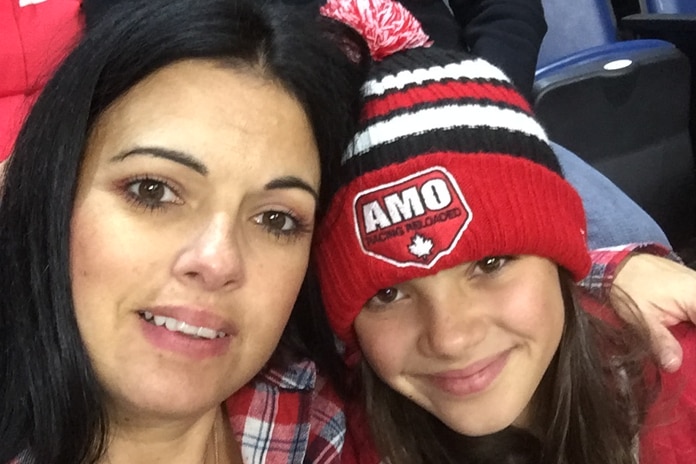 A mother and daughter, wearing a red, black and white beanie, hug and smile.