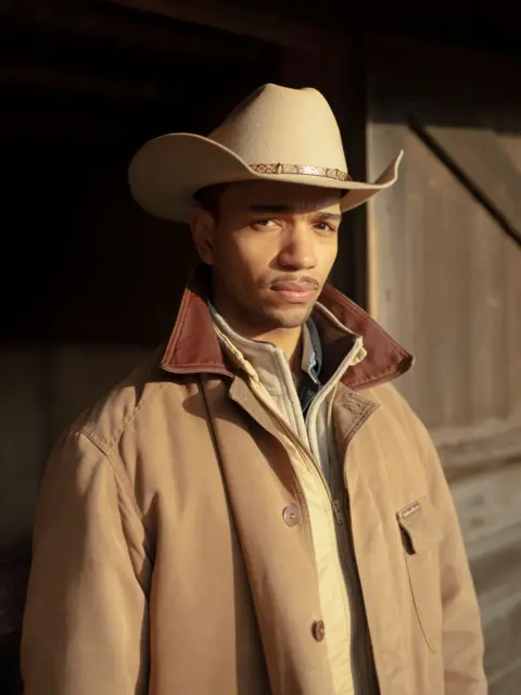 Portrait of Britain/John Boaz Model Benjamin wears a stetson hat, light brown jumper and coat, with a darker collar. He has a moustache. 