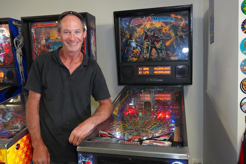 Greg Sharp smiles next to a pinball machine.