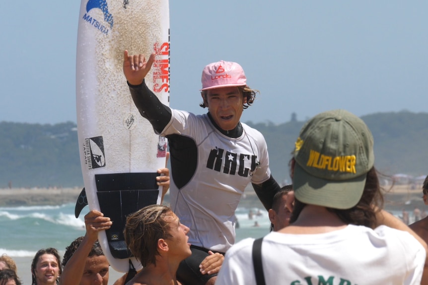 Josh on two men's shoulders with his board behind him. 