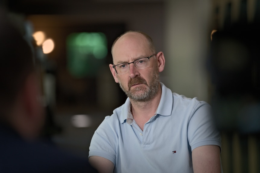A bearded, middle-aged man in glasses wears a polo shirt while sitting in a dimly-lit room.