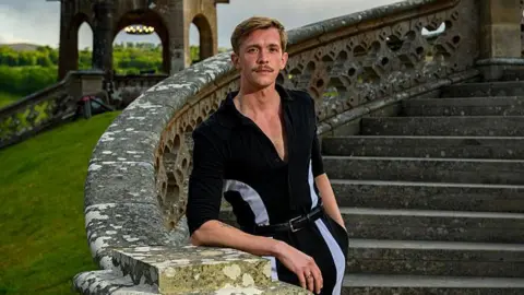 BBC/Studio Lambert/Paul Chappells Stephen Libby, a man with blonde hair and a well groomed moustache standing on a grand outdoor staircase, which he is leaning on with his left arm. He is wearing a striking black jumpsuit with a deep v-neck and white binding down the sides.