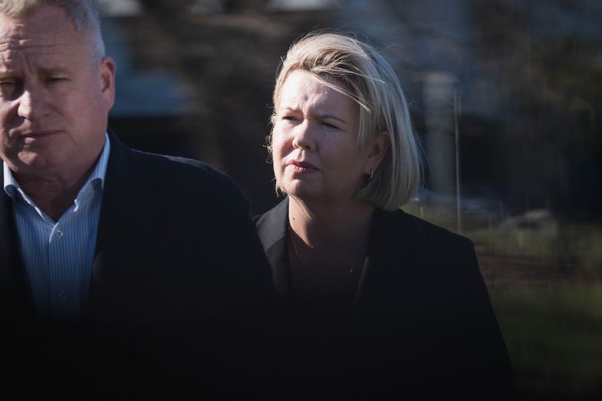 A blonde woman in dark formal wear stands outdoors.
