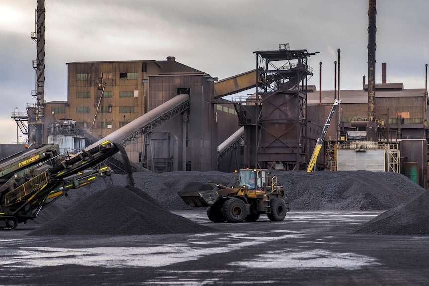 A bulldozer moves black material at a processing facility surrounded by conveyors, large buildings, and chimneys.