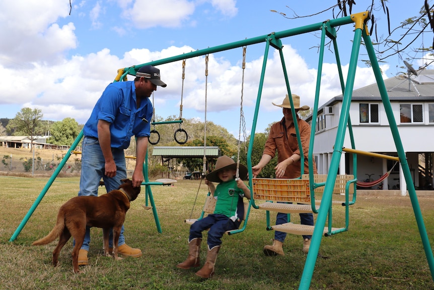 A mum pushing her son on a swing, while the boy's dad stands nearby patting the family dog.