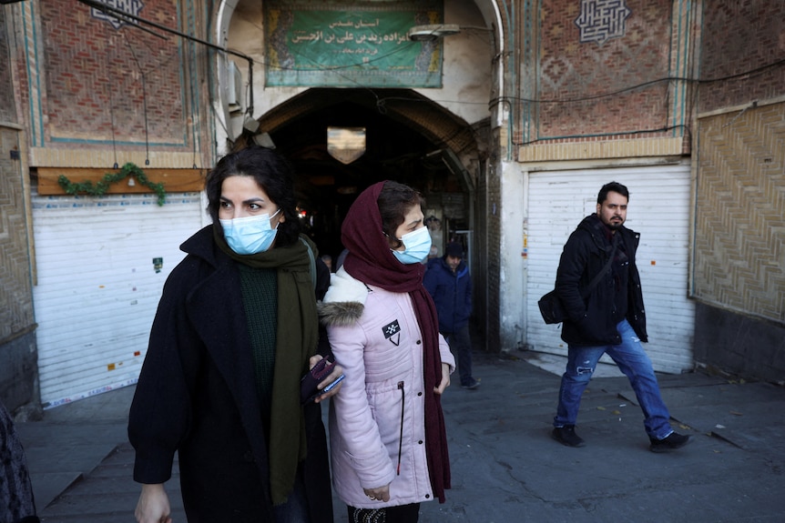 People with masks walking in front of a building with a brick facade and a banner overhead.