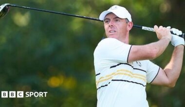 Rory McIlroy watches his ball after hitting a shot with a wood. He is wearing a European Ryder Cup outfit - a baseball cap and a predominantly white polo shirt with black and yellow narrow hoops and trim.