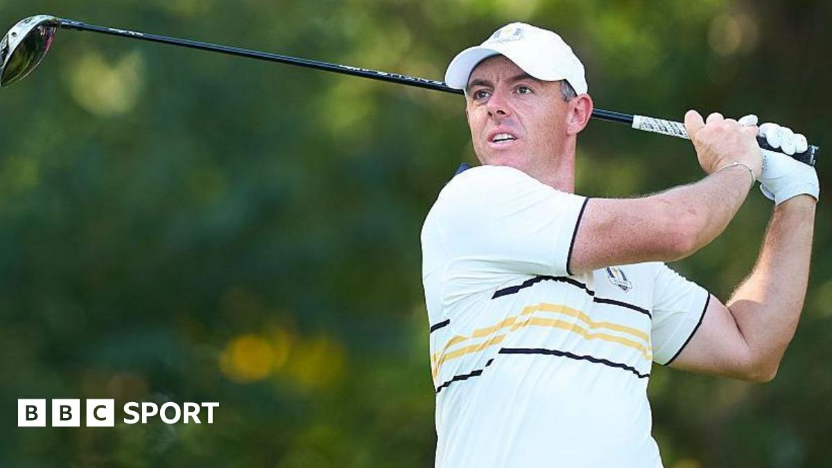Rory McIlroy watches his ball after hitting a shot with a wood. He is wearing a European Ryder Cup outfit - a baseball cap and a predominantly white polo shirt with black and yellow narrow hoops and trim.