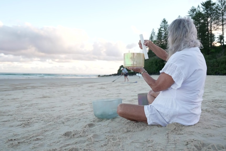 Woman sits crossed legged holding sound bowl on the beach.