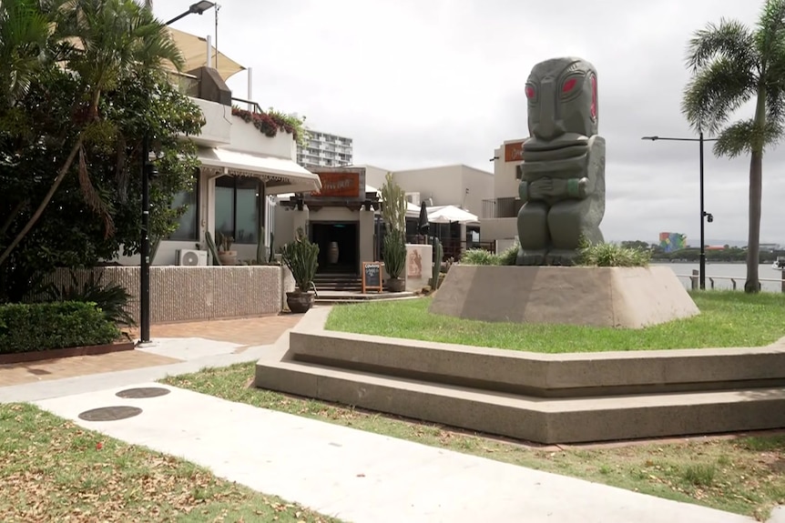 A tiki statue in a public space on the Gold Coast surrounded by bars and water to the right of frame