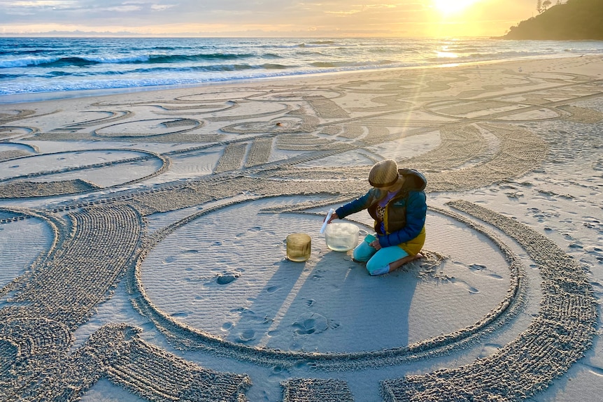 Woman sits in centre of art in the sand with sound bowls.