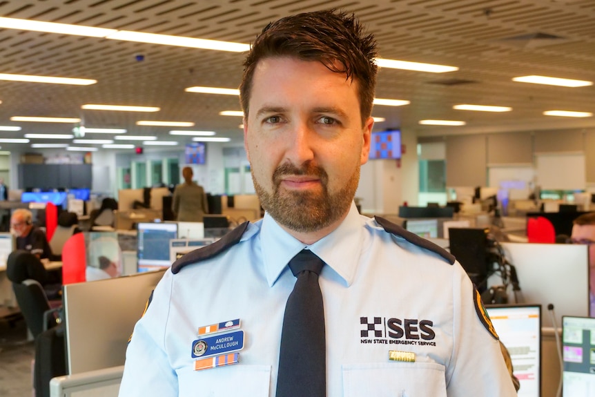 A dark-haired, bearded man in an SES uniform stands in front of workbays in an office.