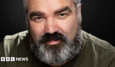 A man with a black and grey beard wearing a khaki green shirt