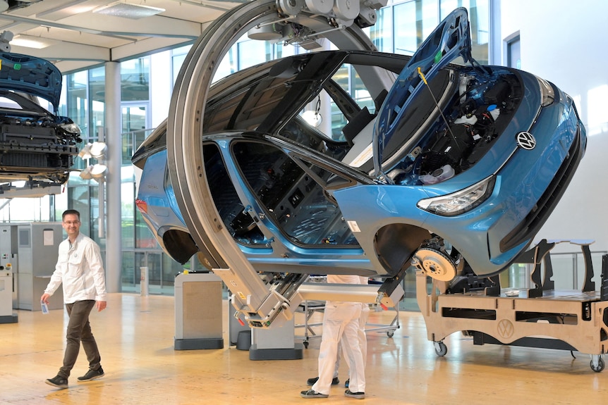 A blue car hangs in a factory as a person works underneath it and another person walks past it