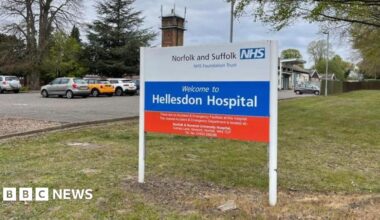 The picture is a close up of the sign for Hellesdon Hospital. It says Norfolk of and Suffolk NHS Foundation Trust  is white red and blue. It also says "Welcome to Hellesdon Hospital". There is a car park behind the sign and some of the cars are visible. The sign it is positioned on grass.