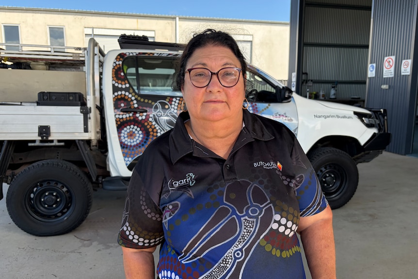A dark-haired woman in glasses and a polo shirt featuring an Indigenous design stands in front of a ute.