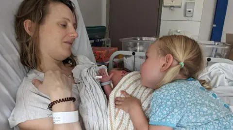 Louise in a hospital bed with her newborn baby and older daughter, who is wearing a blue floral dress and has blonde hair