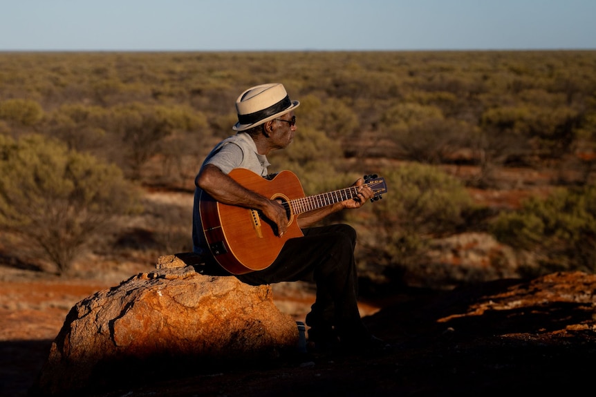 A man wearing a hat sits on a rock playing a guitar on a hill. Beneath the hill tops of green trees can be seen. 