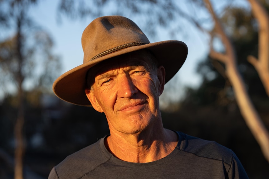 A man wearing a broad rimmed hat stands with his head tilted. He has the glow of sun on his face and trees in the background.