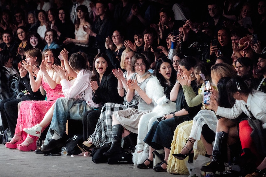 A group of women sit on the edge of a catwalk clapping and cheering as one covers her face in shock.