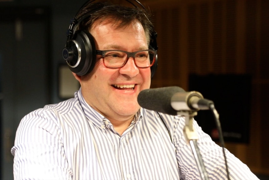 ABC Classic presenter Damien Beaumont stands behind a microphone with over-ear headphones. He is laughing.