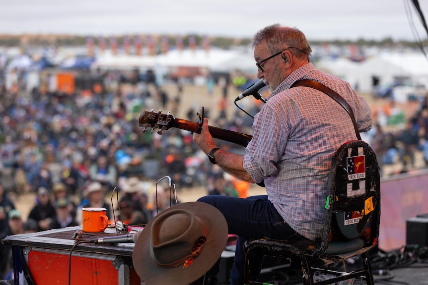 A man plays guitar and signs on stage in front of a large audience. Photo is taking from behind showing man and crowd.