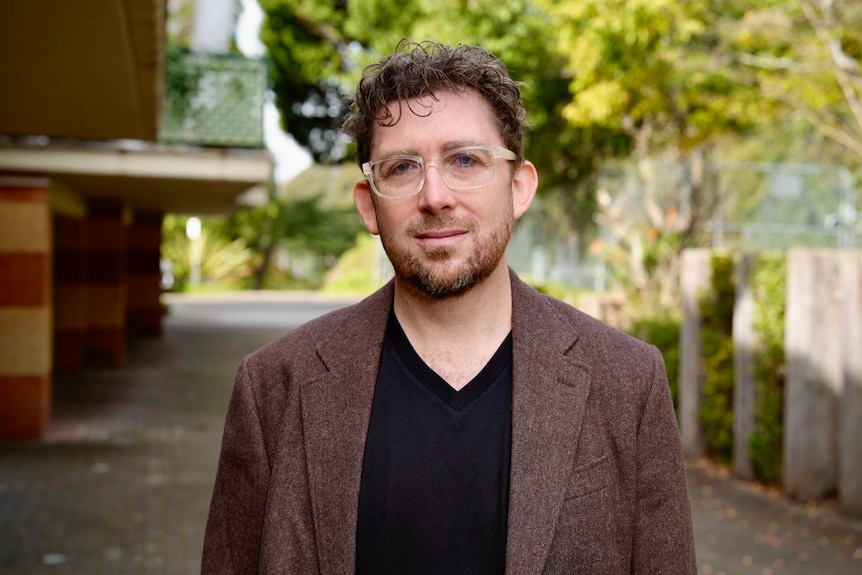 Man with a brown jacket and glasses on looks at camera on the street