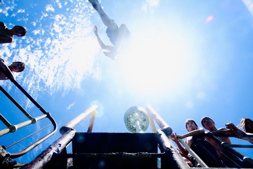 A low shot of a boy jumping off a jetty with his friends clustered on the jetty watching, the sun flares in the blue sky