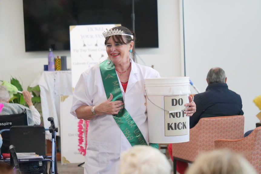 A woman wearing a white coat and pageant sash and crown holding a bucket with "Do Not Kick" printed on it.
