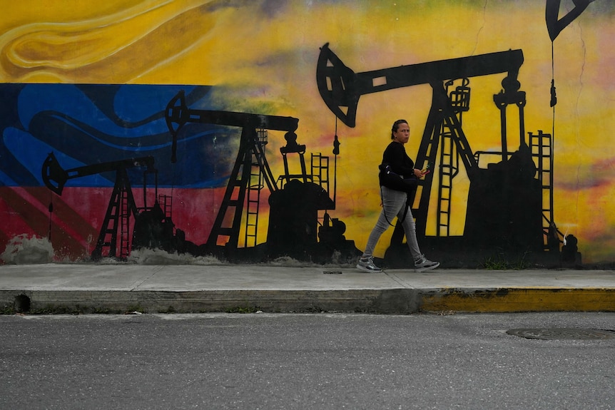 The silhouettes of oil pumps are painted against a yellow background on an outdoor wall, where a woman walks.