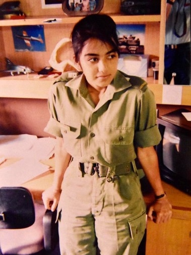 A woman wearing a military uniform leans on a table and looks on.