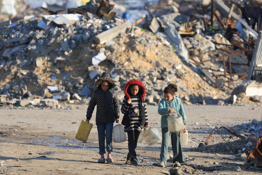 Three young girls walk with buckets in their hands with a background of rubble