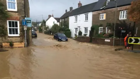 Steve Smith A residential street is heavily flooded, with muddy water flowing past rows of houses. Several cars are partially submerged, and a person in high‑visibility clothing stands near the roadside as the floodwater stretches across the entire road.