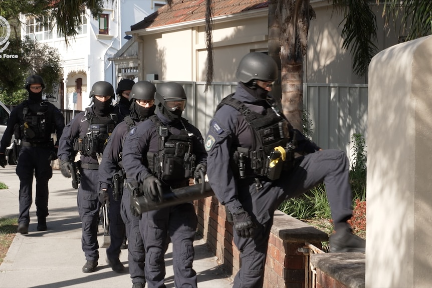 A group of riot squad officers in dark uniforms and padded vests, preparing to force entry into a home.