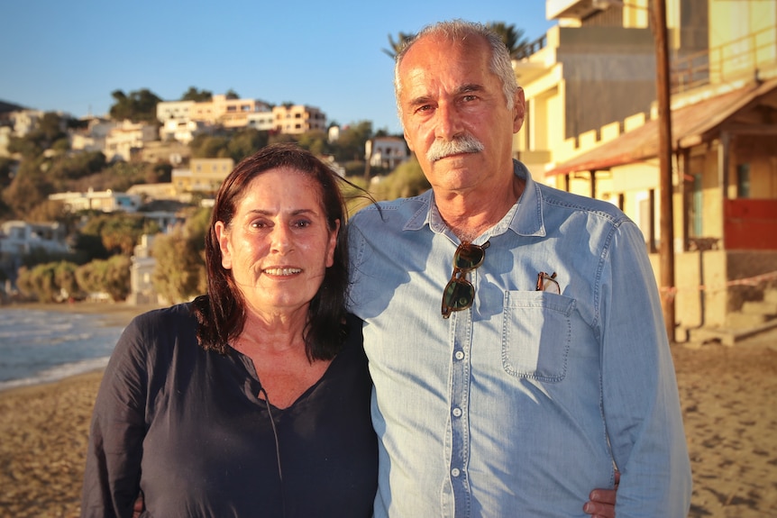 Woman and man standing closely together on a beach, Greek island behind them. Him in blue shirt, her in dark blue top.