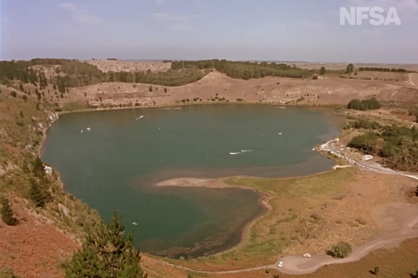 An old restored photo of a large blue-green lake surrounded by steep crater edges with trees and grass on them.
