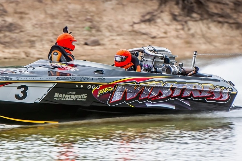 Two men wearing orange helmets in a black power boat, speeding along a river