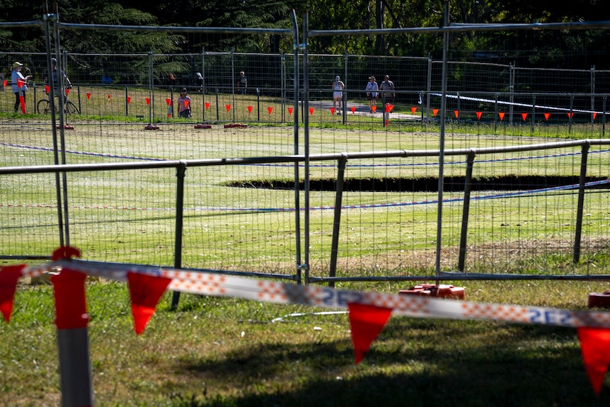 A large hole in a field of green grass is fenced off with temporary fencing and SES tape with orange flags.