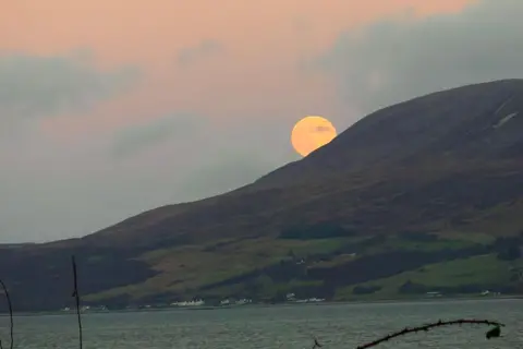 BBC Weather Watchers/TealKat A yellow/pink moon peaks out from behind a dark green hill on a low incline. There is a loch in front of the hill and it is dusk, so the sky is orangey pink