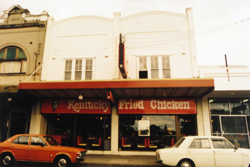 A two-storey building with a red verandah and Kentucky Fried Chicken signage with red and white tables and chairs inside.