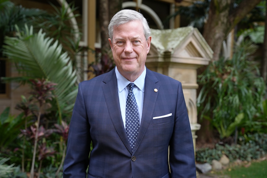 A man in a suit and tie standing in front of a garden
