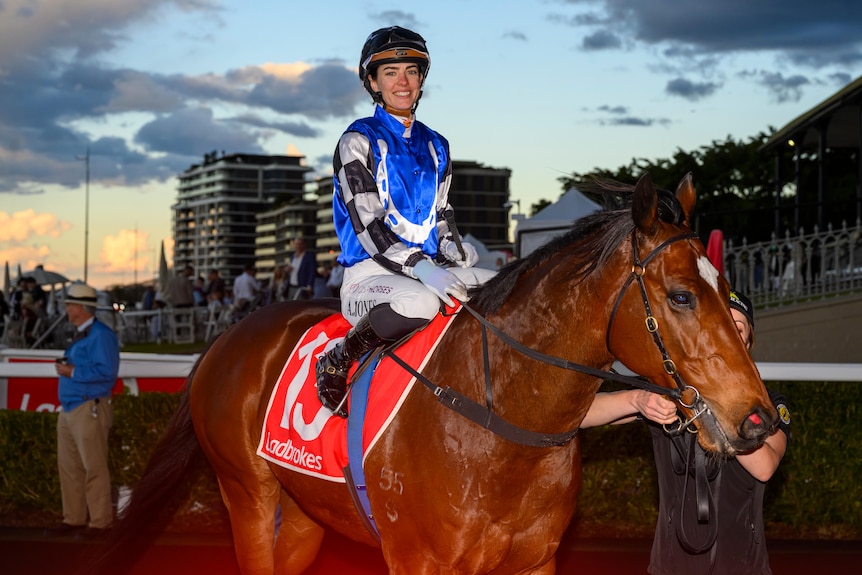 A young female jockey wearing blue, black and white silks sitting on a horse.