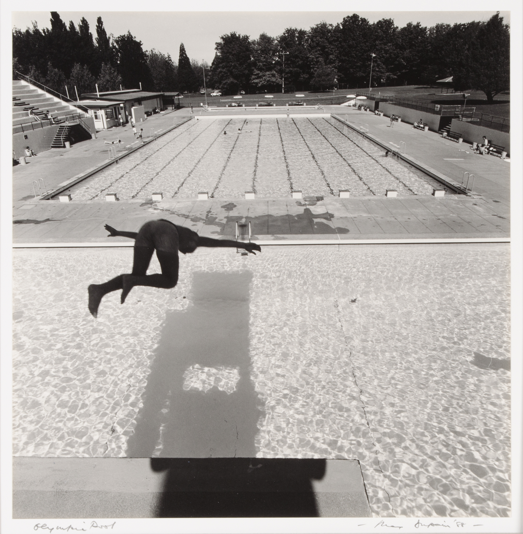A black and white photograph of a shadowed figure diving from a diving tower into a pool, an Olympic-sized pool beyond.