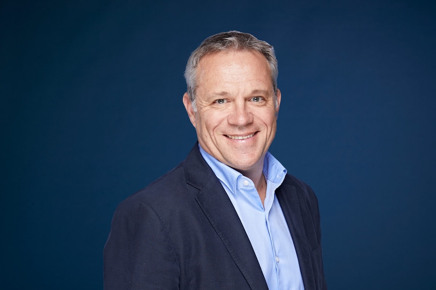 A 50-something man in a blue shirt and dark suit smiles at camera in a portrait shot