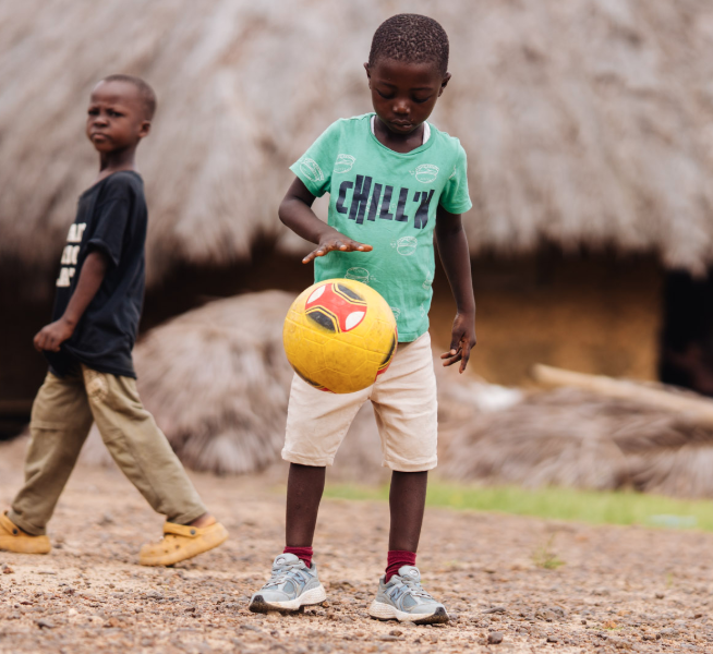 A boy in a green T-shirt bounces a soccer ball with a palm roofed hut behind him