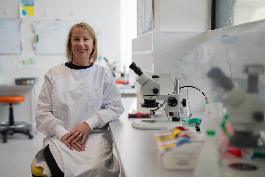 A lady in a lab coat in a lab, smiling at the camera.