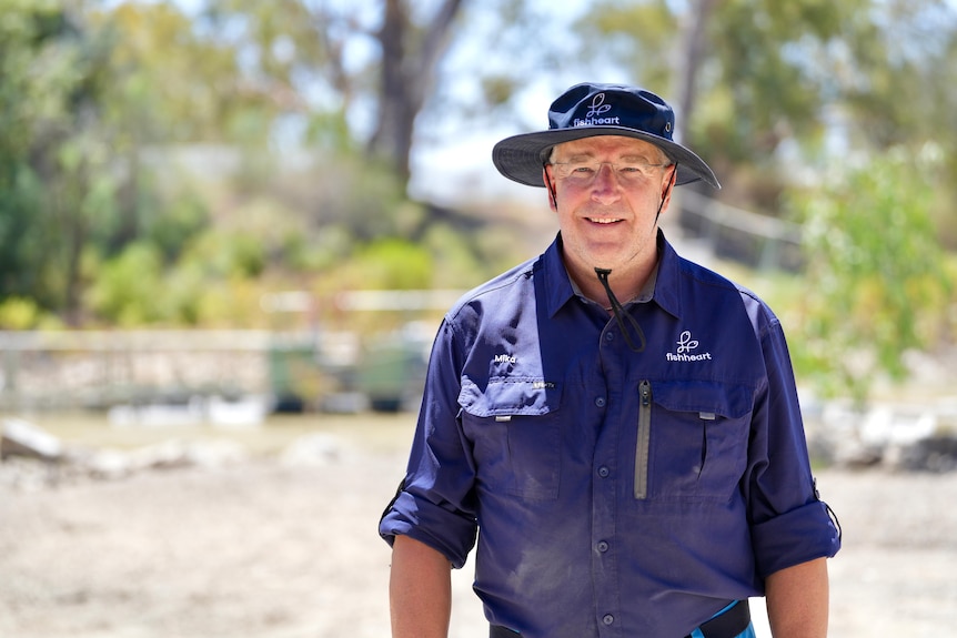 A white man wearing a blue shirt and hat which read Fishheart standing on the banks of the Darling River